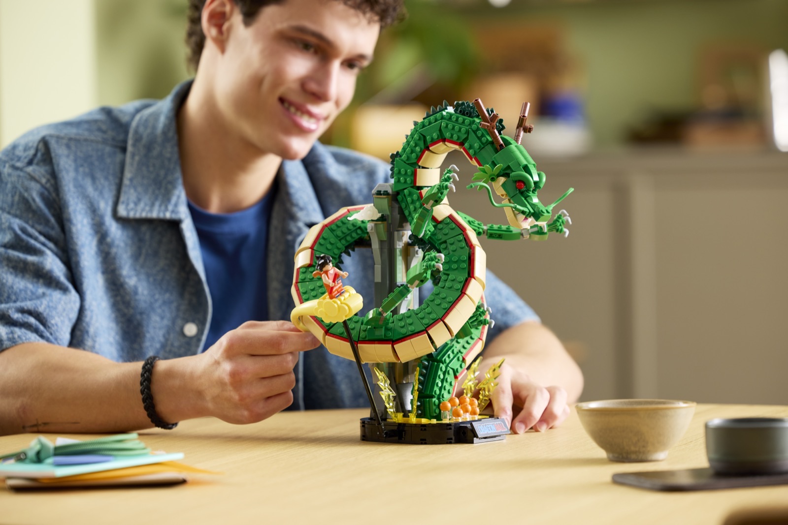Ein junger Mann sitzt an einem Holztisch und rückt die Figur Goku auf ihrer gelben Wolke neben dem grünen LEGO Drachen Shenron zurecht