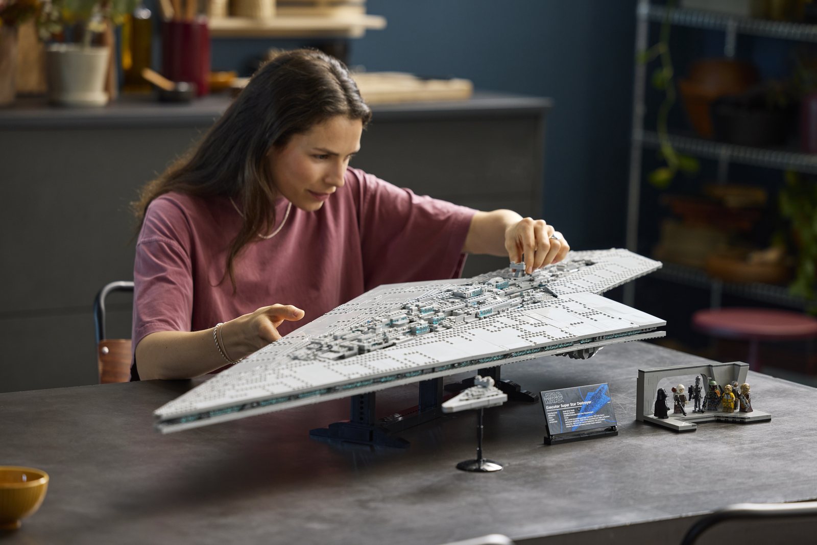 Eine Person setzt am Esstisch ein Bauteil auf den Rumpf des fertigen LEGO Supersternzerstörers, daneben der kleine Sternzerstörer, die Infotafel und die Brückenszene