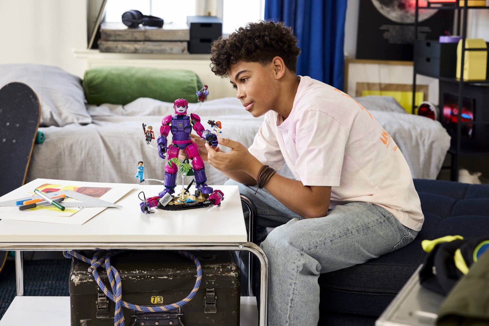 Ein Jugendlicher richtet am Schreibtisch die Minifiguren am lila-pinken Sentinel-Roboter aus dem LEGO-Set Legendärer Showdown 76348 aus, mehrere Figuren schweben an transparenten Stäben um den Roboter
