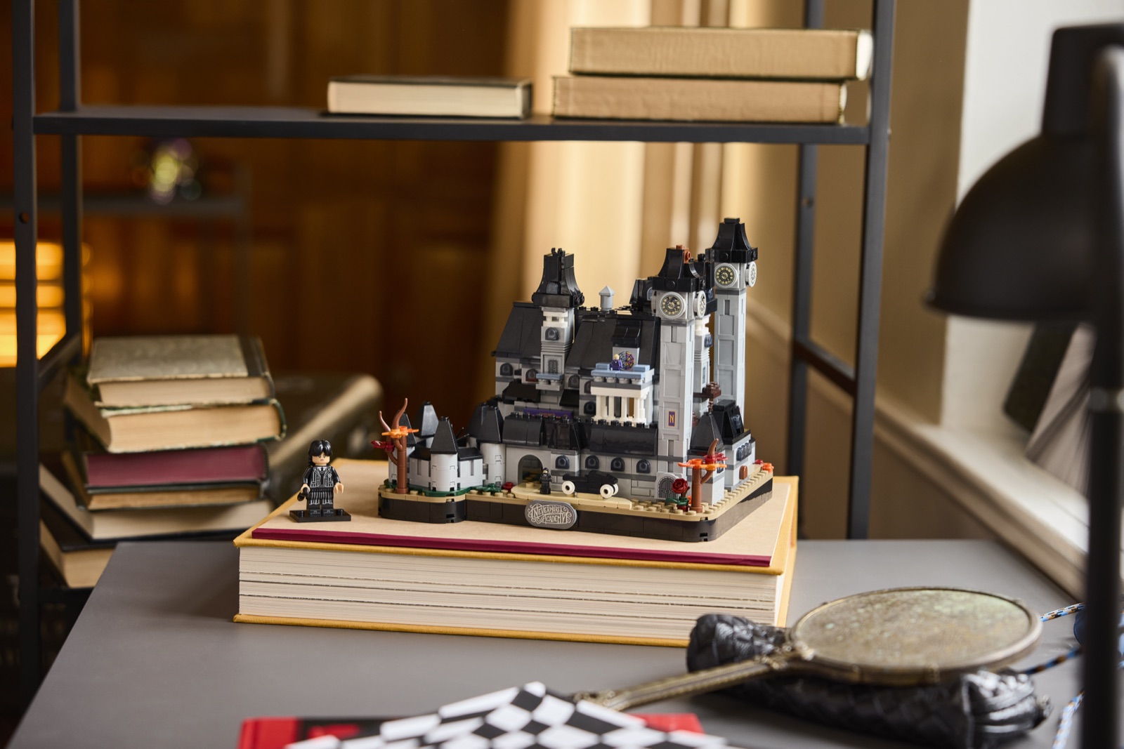Die Nevermore Academy aus LEGO-Steinen im Mikromaßstab steht auf einem großen Buch auf dem Schreibtisch, links daneben die Wednesday-Minifigur