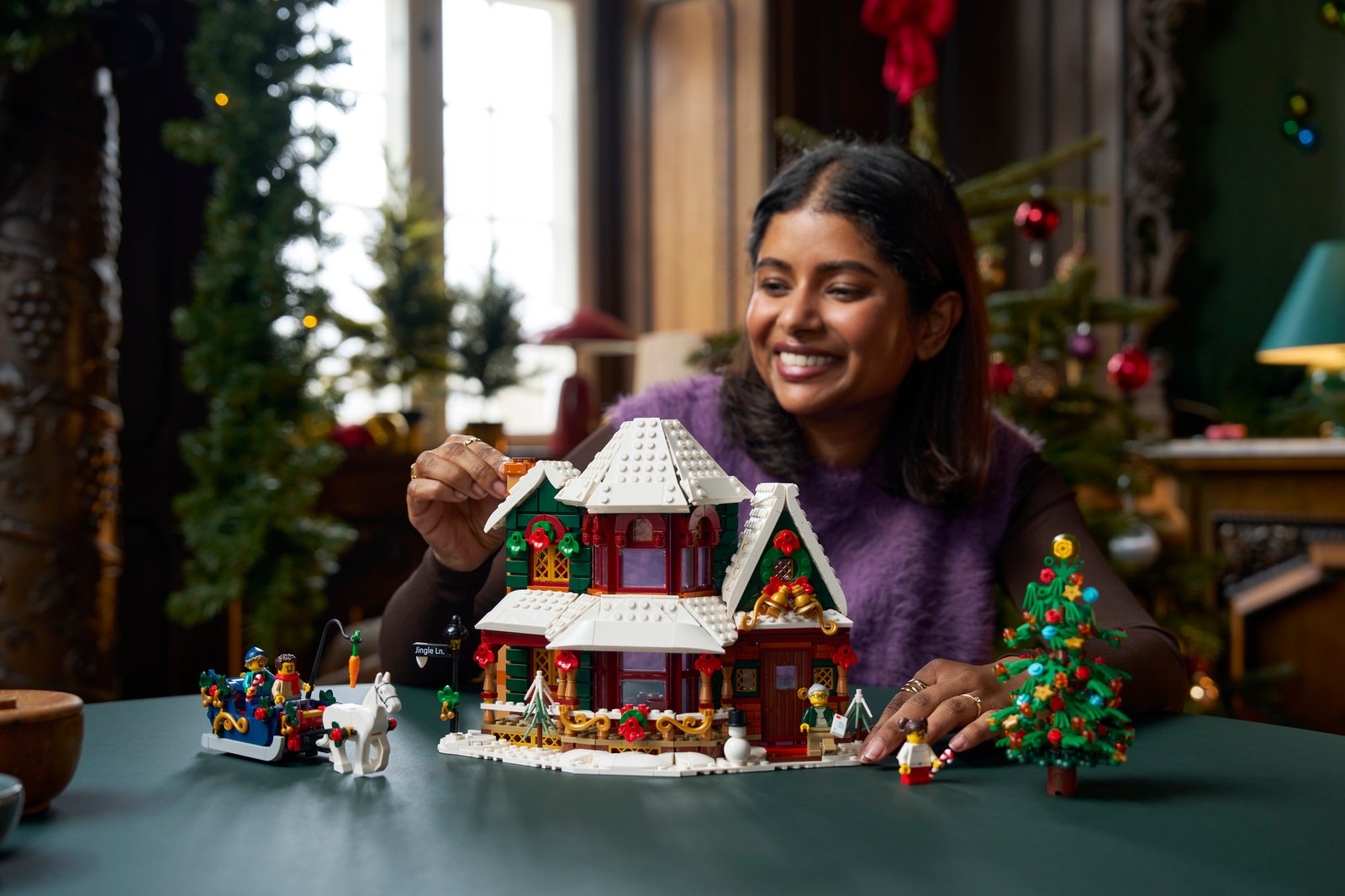 Eine Frau baut am LEGO Weihnachtshaus 11387 mit verschneitem Dach, daneben stehen der Pferdeschlitten, ein geschmückter Weihnachtsbaum und Minifiguren