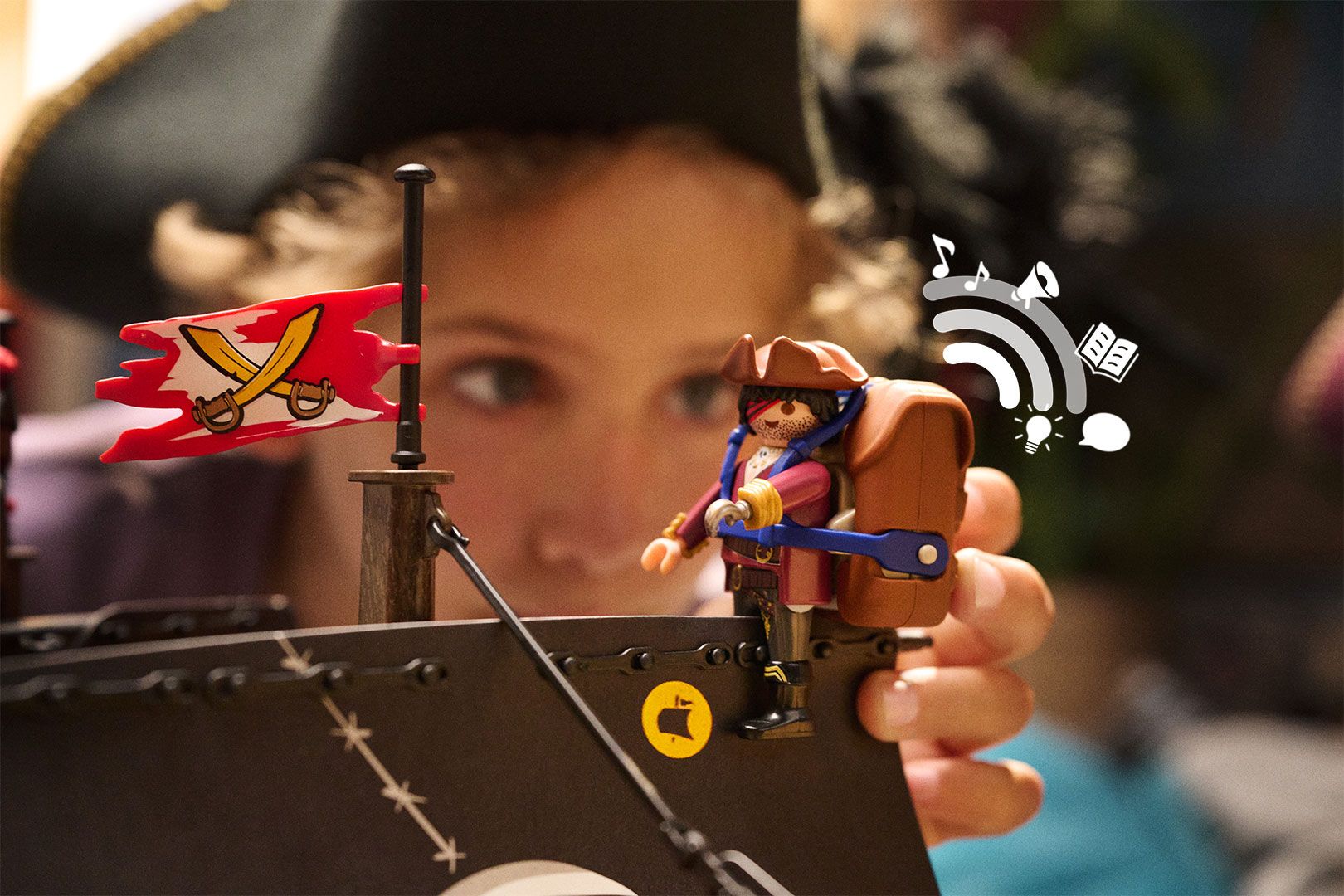 Ein Kind hält eine Playmobil-Piratenfigur mit braunem Audio-Rucksack an den Bug eines Piratenschiffs
