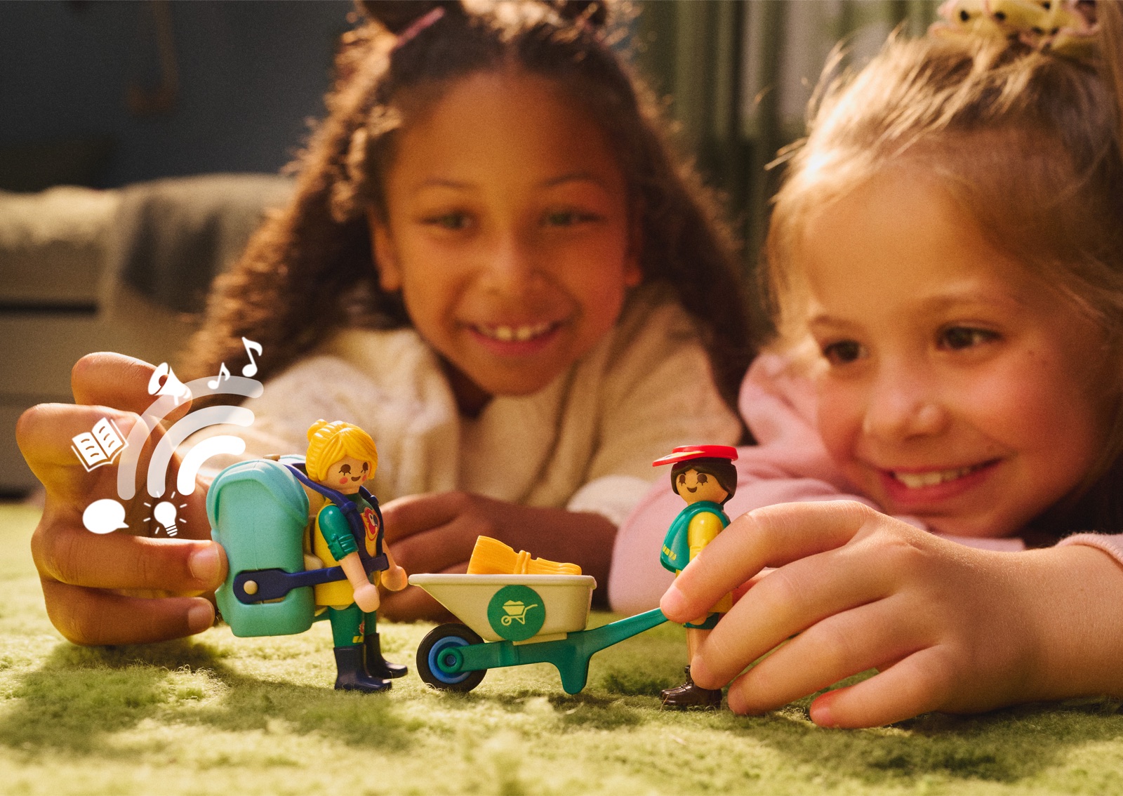 Zwei Kinder spielen auf einem Teppich mit Playmobil-Zoofiguren, eine Tierpflegerin trägt einen grünen Audio-Rucksack